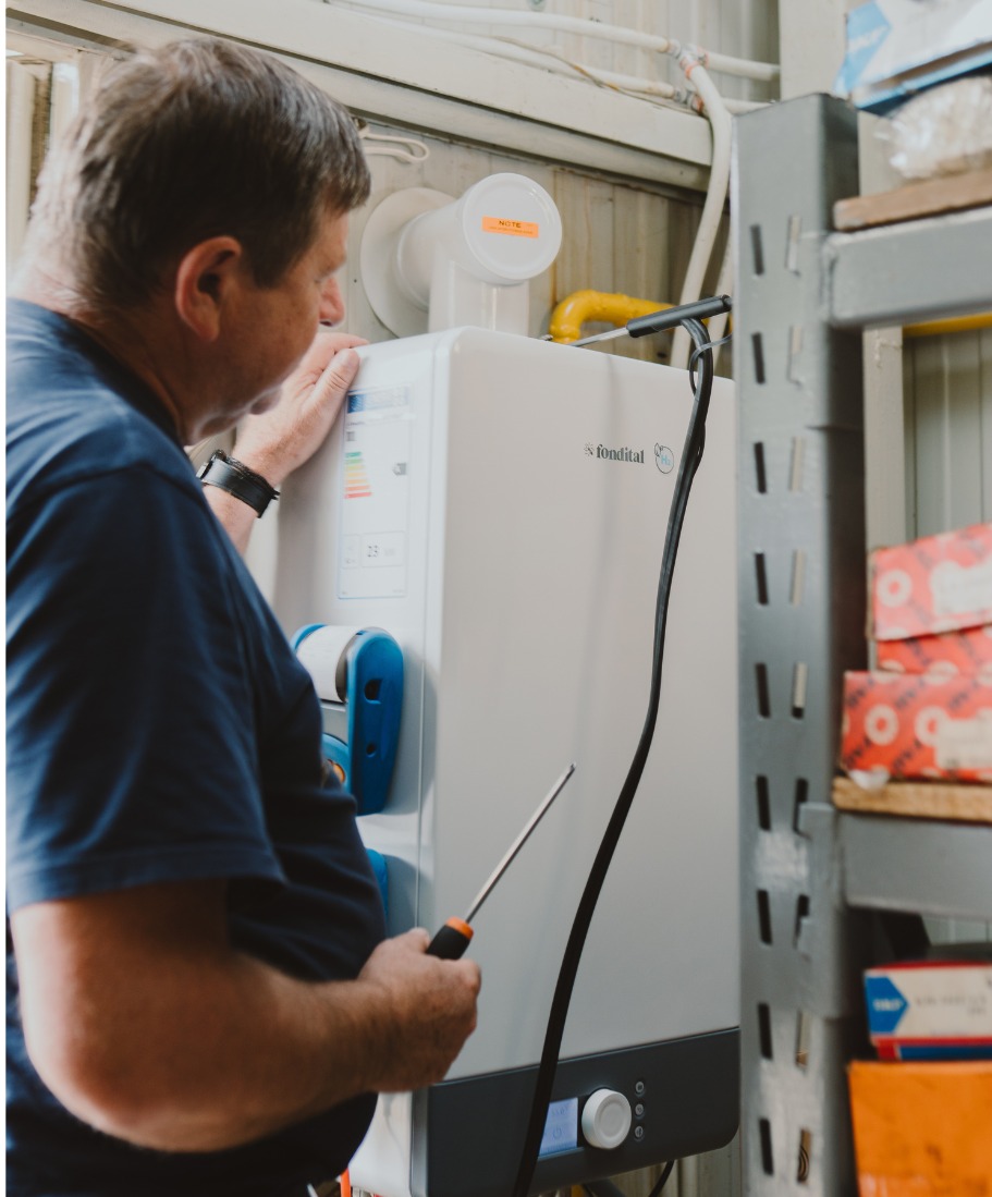 Mężczyzna w niebieskiej koszulce serwisuje biały, wiszący piec gazowy marki Fondital, używając śrubokręta. Widoczne rury, półki z pudełkami i naklejka z napisem.
