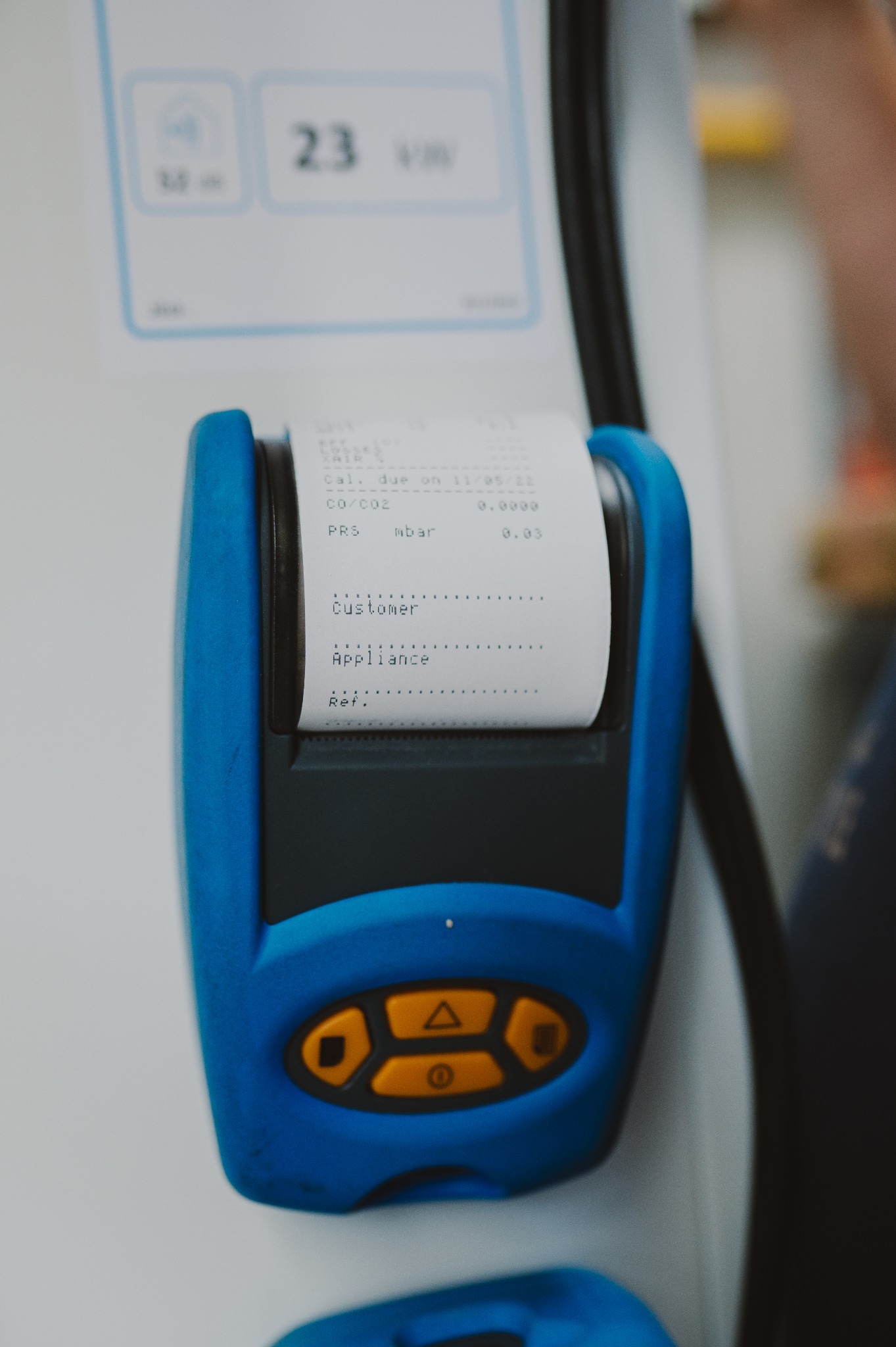 Niebieski analizator spalin z wydrukiem parametrów CO/CO2 i ciśnienia, datą kalibracji na tle urządzenia pomiarowego i fragmentu osoby.