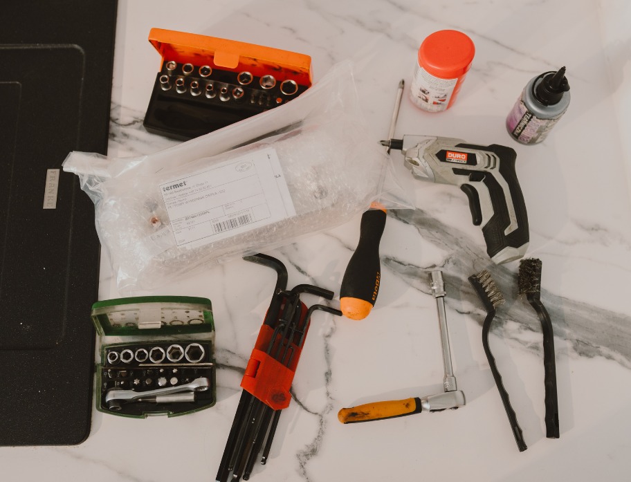 Zestaw narzędzi hydraulicznych i akcesoriów (wiertarka, klucze, szczotki, nasadki) rozłożony na marmurowym blacie. Widoczna paczka z elementem instalacji Termet.