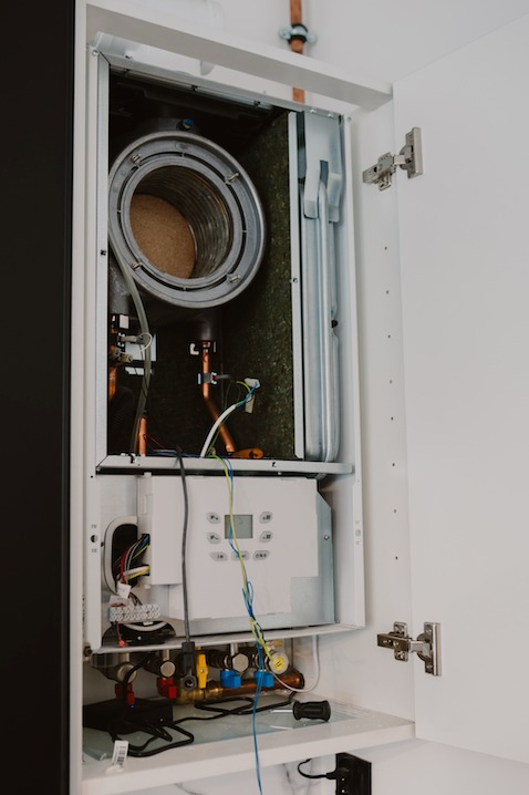 Otwarty piec gazowy w białej szafce, widoczne elementy instalacji i narzędzia. Miedziane rury i kable wskazują na trwający serwis lub naprawę.