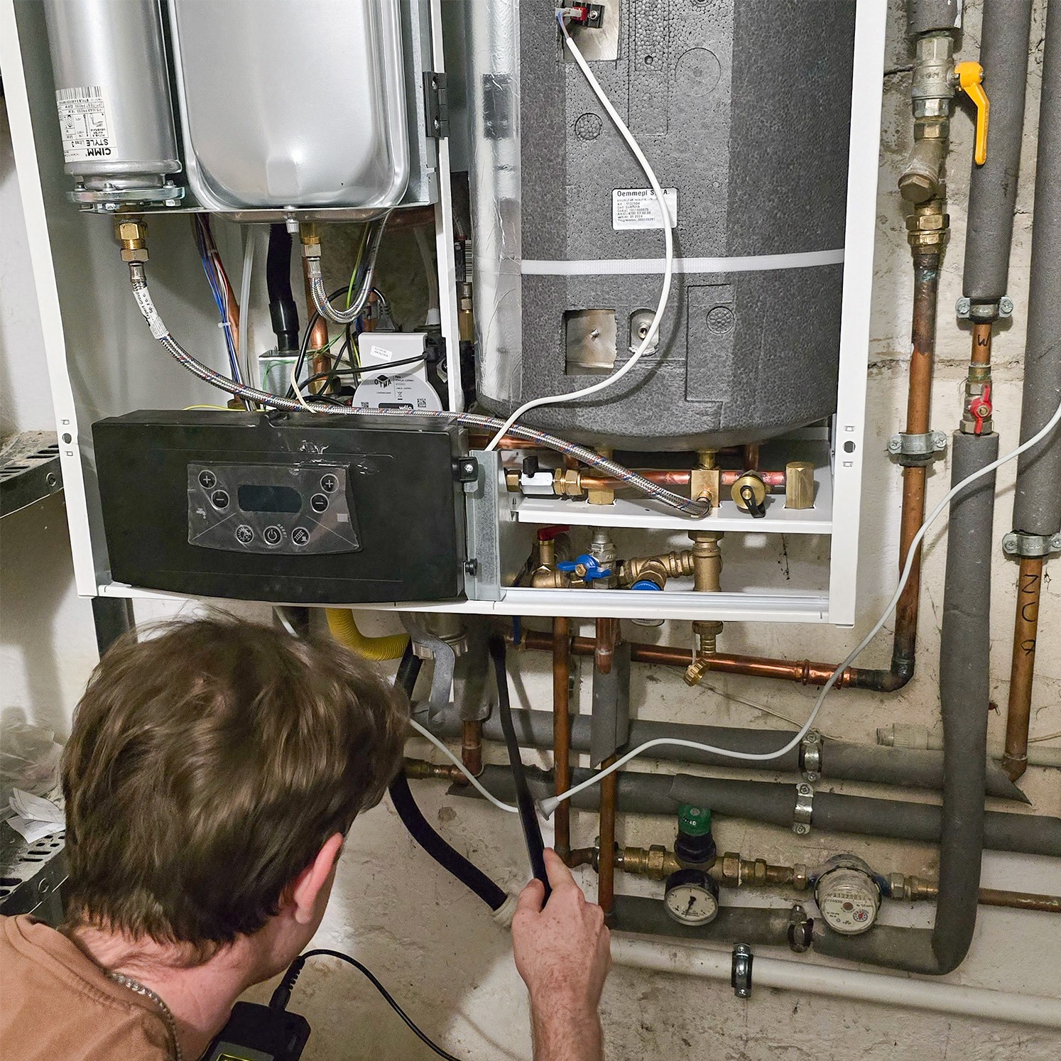 Mężczyzna kontroluje piec gazowy za pomocą urządzenia diagnostycznego, widoczne rury, zawory i elementy instalacji.