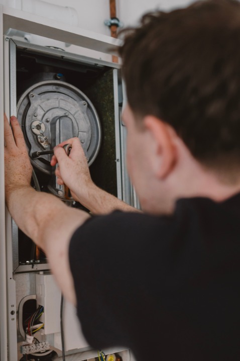 Serwisant w czarnej koszulce naprawia piec gazowy. Widoczne detale wnętrza pieca i narzędzia w rękach. Ujęcie zza pleców serwisanta.