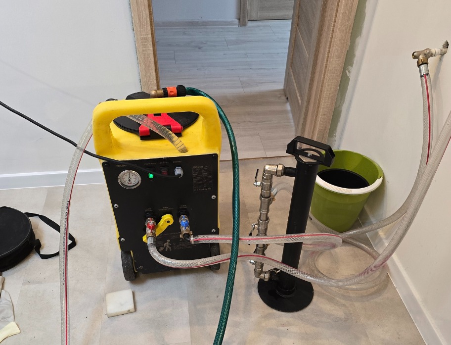 Żółte urządzenie do przepychania rur podłączone do instalacji, z widocznymi przewodami i manometrem, w pomieszczeniu z otwartymi drzwiami w tle.