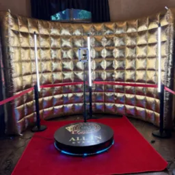 Złota ścianka i platforma obrotowa ze światłami LED na czerwonym dywanie, tworzące wystawne tło do zdjęć.