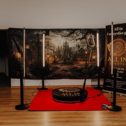 All in events - Dekoracja Halloween z fotobudką 360, tło z motywem zamku i dyni. Czarny pająk na ścianie. Podest fotobudki na czerwonym dywanie. Atmosfera imprezy.