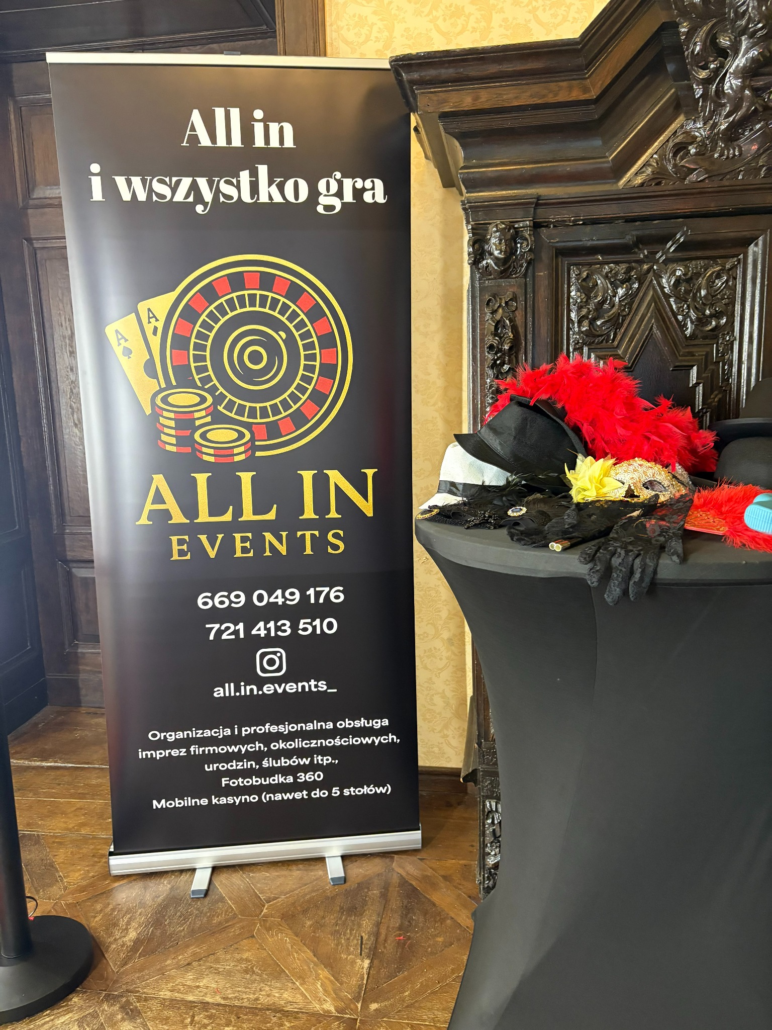 Roll-up z logo 'All In Events' i grafiką kasyna, obok stolik z rekwizytami: kapelusze, maski, boa. Dekoracje na imprezę tematyczną w eleganckim wnętrzu.