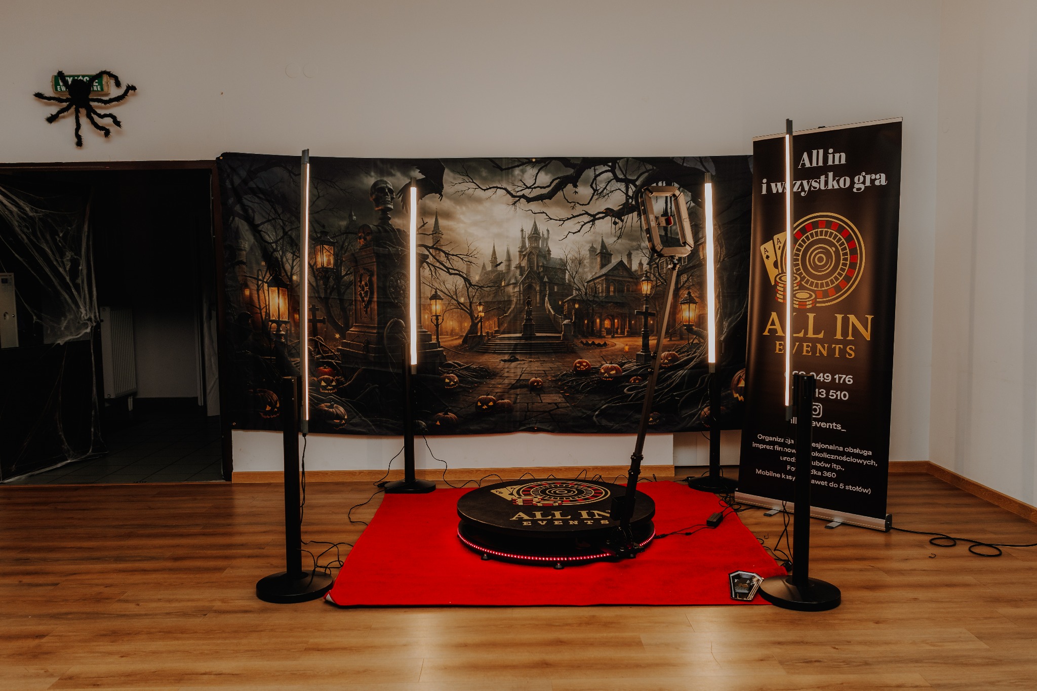 Dekoracja Halloween z fotobudką 360, tło z motywem zamku i dyni. Czarny pająk na ścianie. Podest fotobudki na czerwonym dywanie. Atmosfera imprezy.