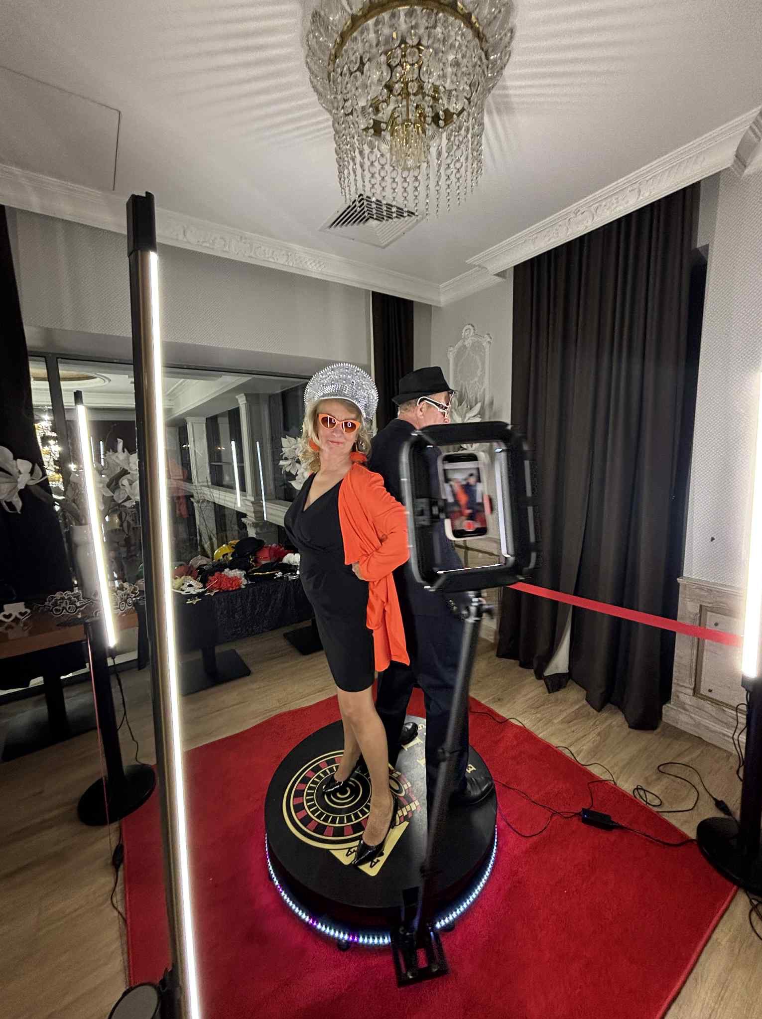 Para w stylizowanych strojach pozuje na obrotowej platformie fotobudki 360 z oświetleniem LED, na czerwonym dywanie w eleganckim wnętrzu z żyrandolem.