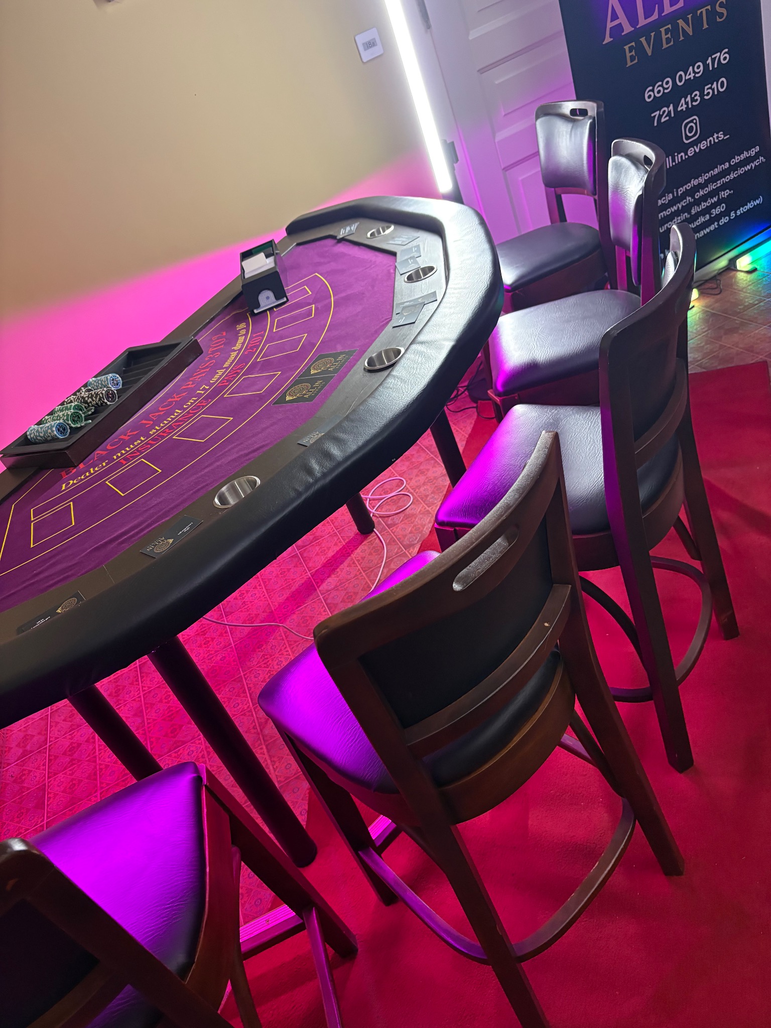 Stół do blackjacka z żetonami i krzesłami, oświetlony różowym światłem, na czerwonym dywanie. W tle baner reklamowy firmy eventowej.