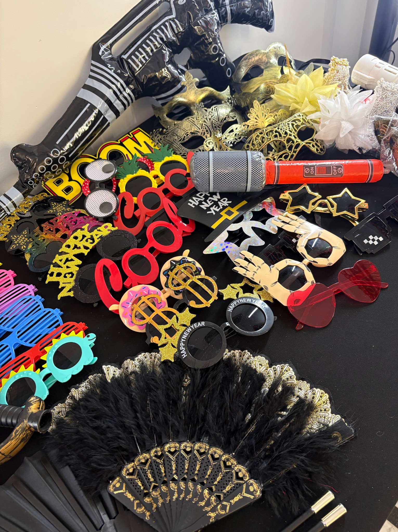 Kolorowe rekwizyty do fotobudki: maski karnawałowe, okulary w kształcie serc, mikrofon, dmuchana gitara i wachlarz z piór na czarnym tle. Akcesoria imprezowe.