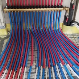 MSB-INSTAL - Rozdzielacz z rurami ogrzewania podłogowego w kolorach niebieskim i czerwonym, ułożonymi na izolacji z folią w kratkę. Widoczne elementy instalacji.