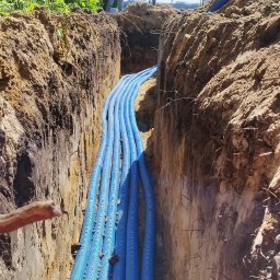 MSB-INSTAL - Niebieskie rury karbowane w wykopie ziemnym, prawdopodobnie instalacja pod ogrzewanie podłogowe. W tle osoba w kasku i słup elektryczny.