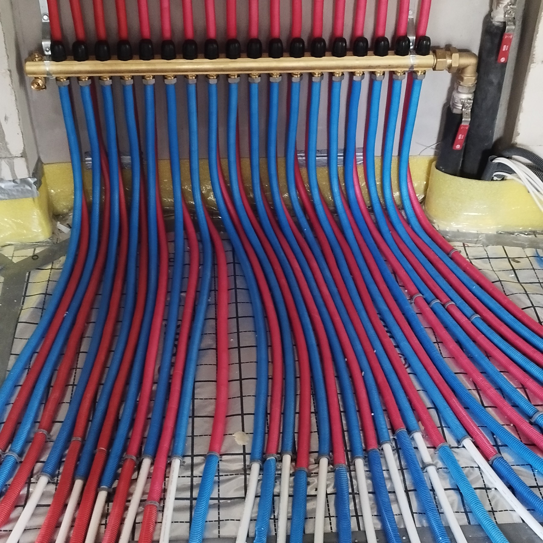 Rozdzielacz z rurami ogrzewania podłogowego w kolorach niebieskim i czerwonym, ułożonymi na izolacji z folią w kratkę. Widoczne elementy instalacji.