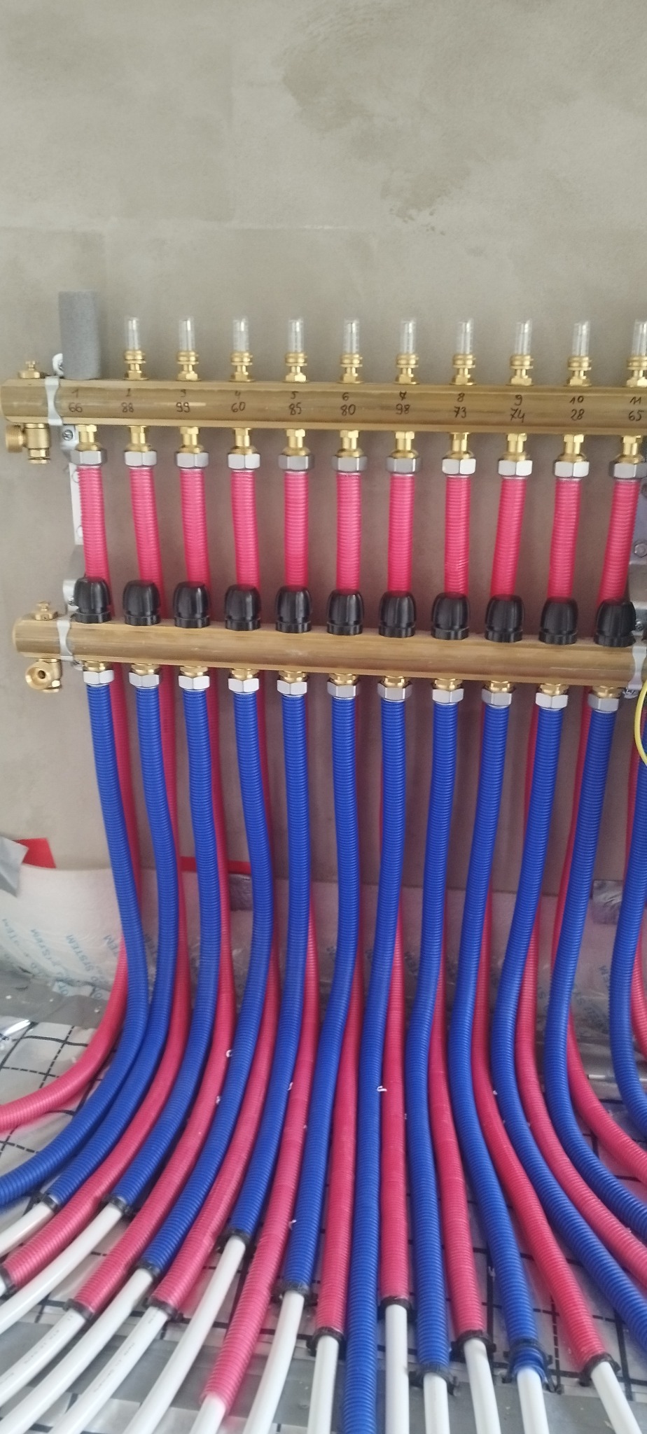 Rozdzielacz ogrzewania podłogowego z rurami w kolorach niebieskim i czerwonym. Widoczne oznaczenia numeryczne na rozdzielaczu. Instalacja w trakcie montażu.