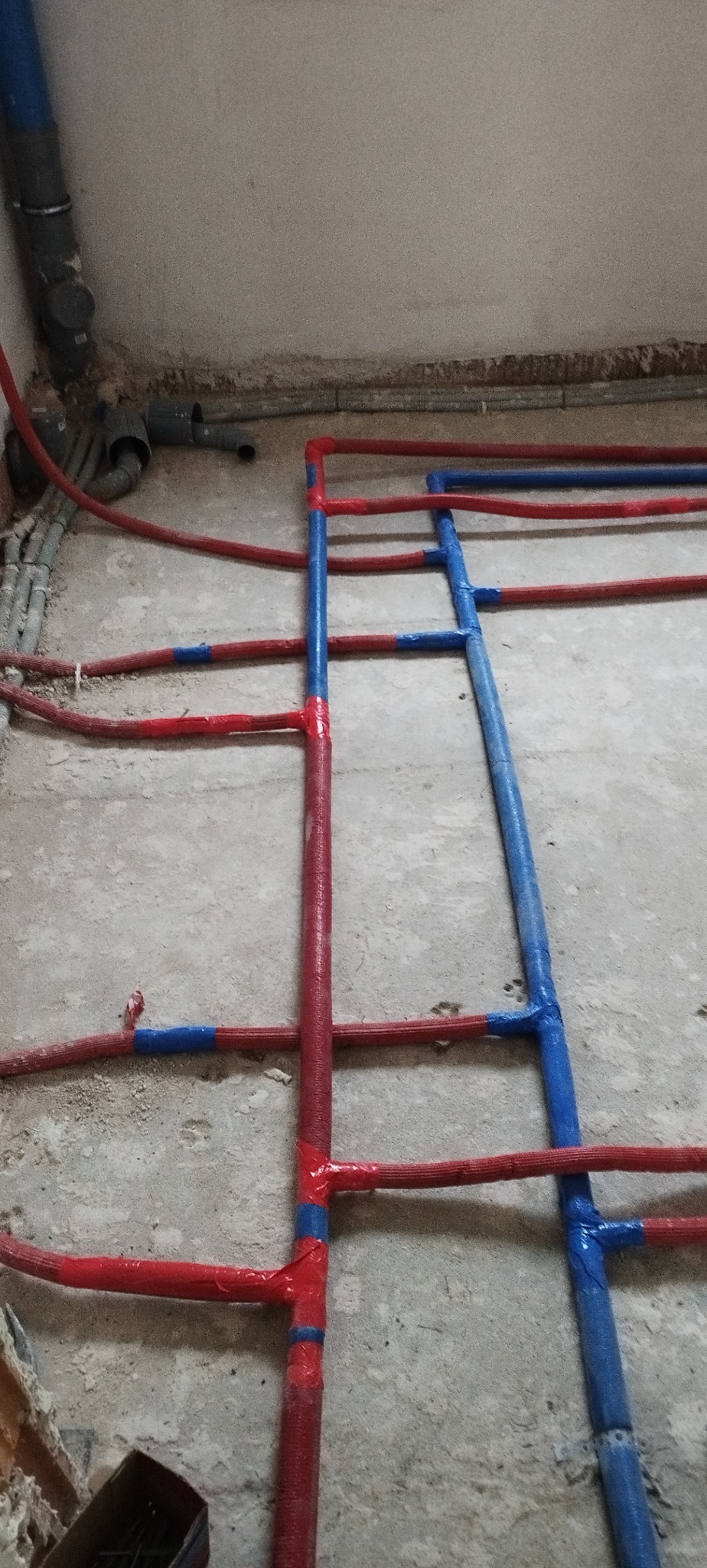 Instalacja grzewcza podłogowa w trakcie montażu, rury w izolacji czerwonej i niebieskiej, widoczne złącza i mocowania na surowym podłożu.