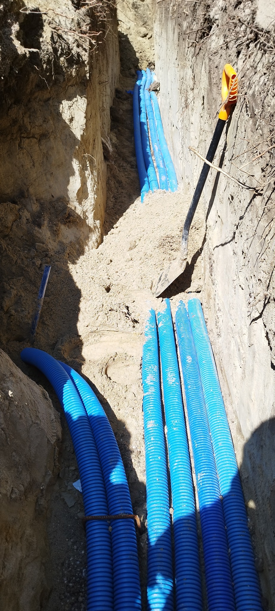 Błękitne rury w wykopie ziemnym, częściowo zasypane piaskiem, z widoczną łopatą. Instalacja podziemna w trakcie realizacji.