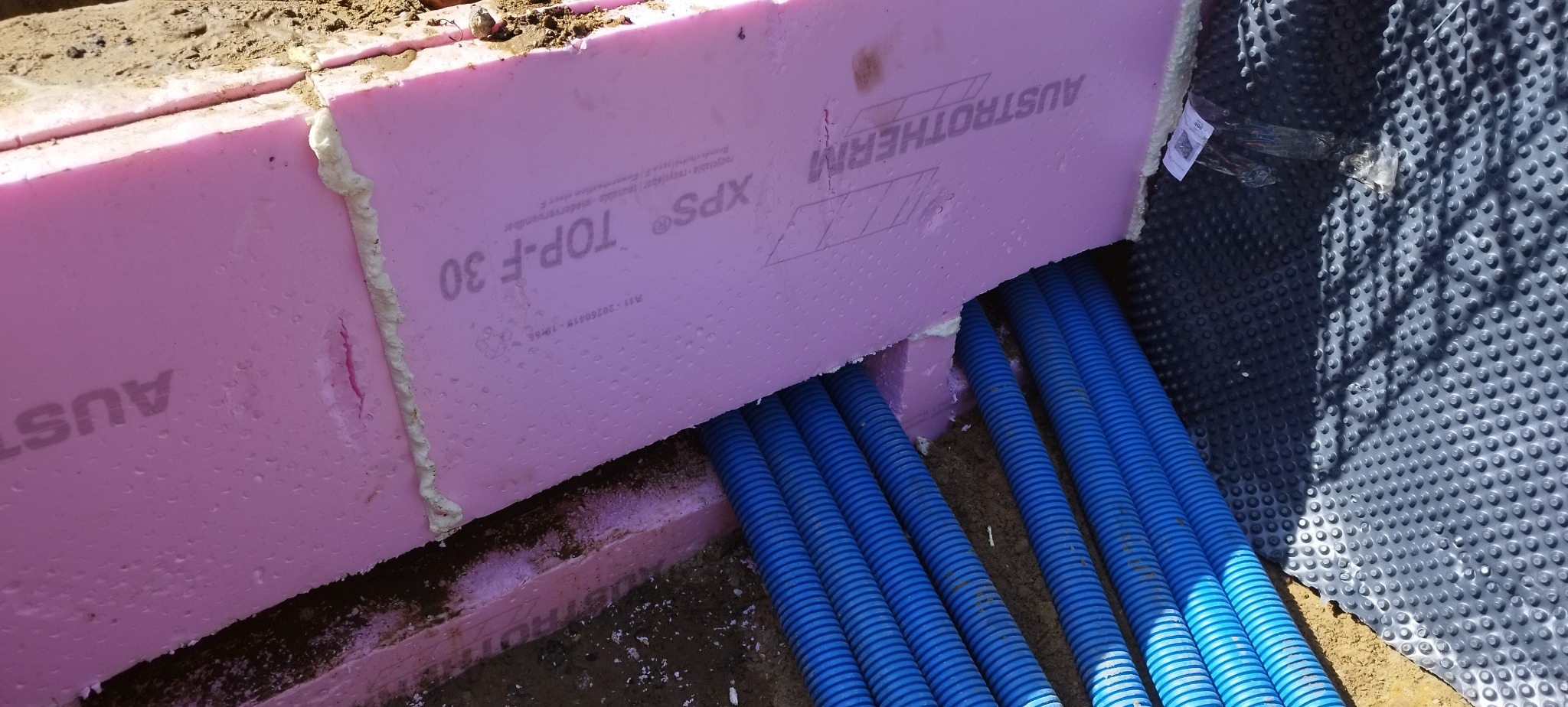 Niebieskie rury w izolacji z różowego styropianu Austrotherm XPS TOP-F 30, widoczne na tle folii kubełkowej, prawdopodobnie ogrzewanie podłogowe.