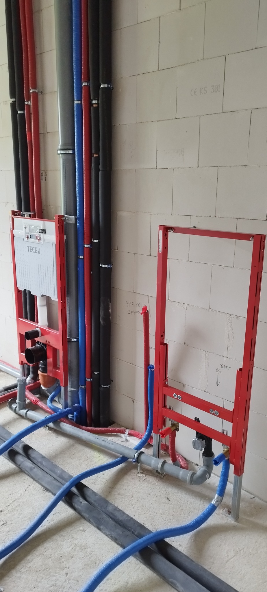 Instalacja wodno-kanalizacyjna w surowym stanie: rury w izolacji (czerwone, niebieskie, czarne) prowadzone wzdłuż ściany z betonu komórkowego, stelaże podtynkowe WC.