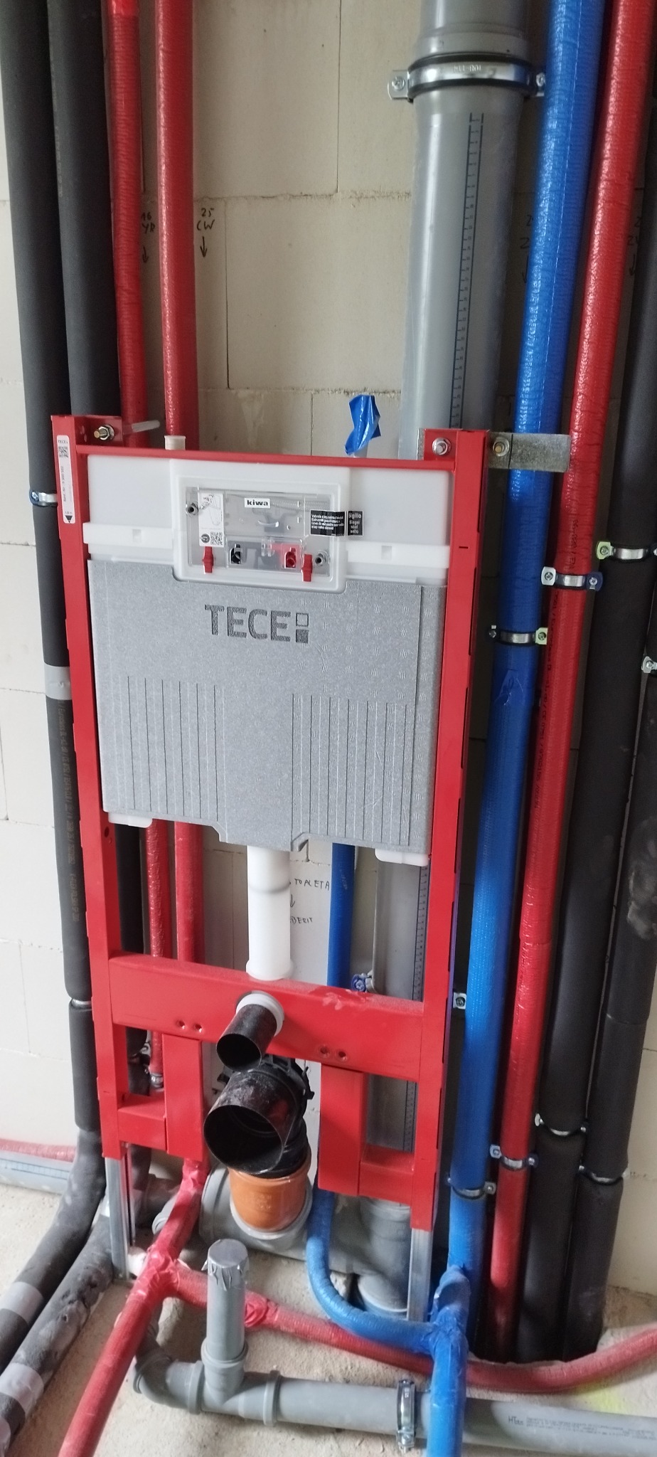 Stelaż podtynkowy WC marki Tece, z widocznymi rurami instalacji wodnej (czerwone, niebieskie) i kanalizacyjnej (czarne, szare) w surowym wnętrzu łazienki.