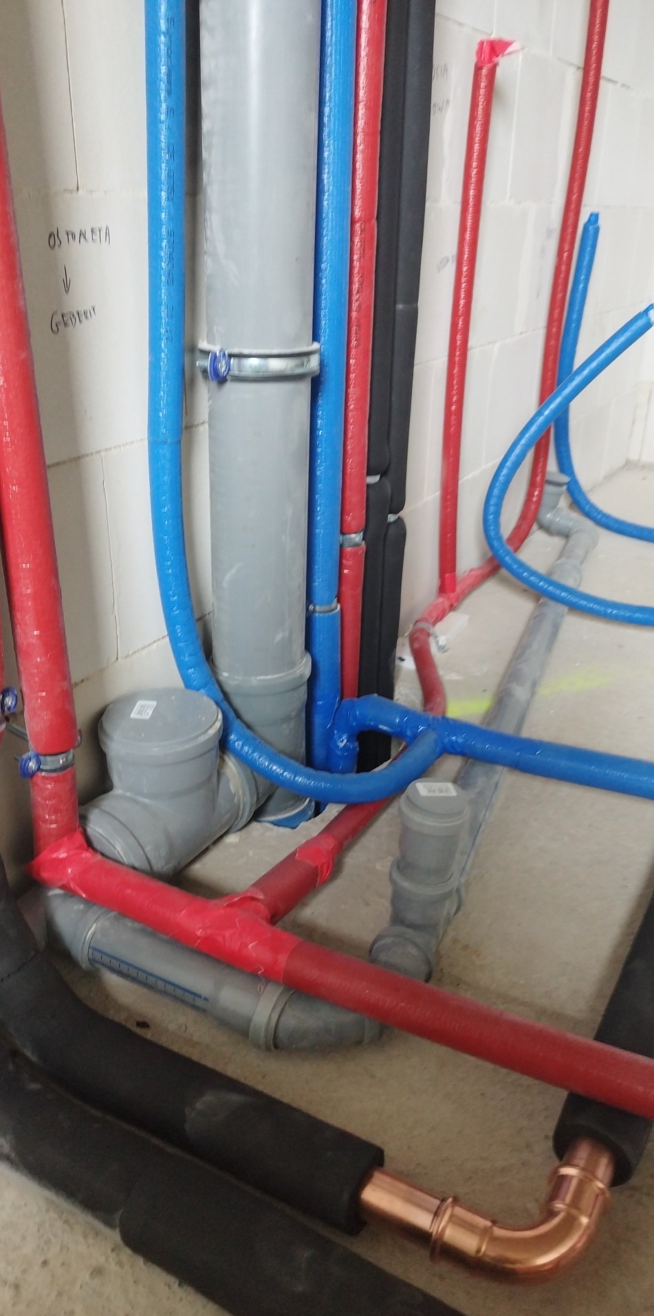 Instalacja wodno-kanalizacyjna w surowym stanie: rury PCV szare, czerwone i niebieskie, niektóre zaizolowane, prowadzone po podłodze i wzdłuż ściany z bloczków.