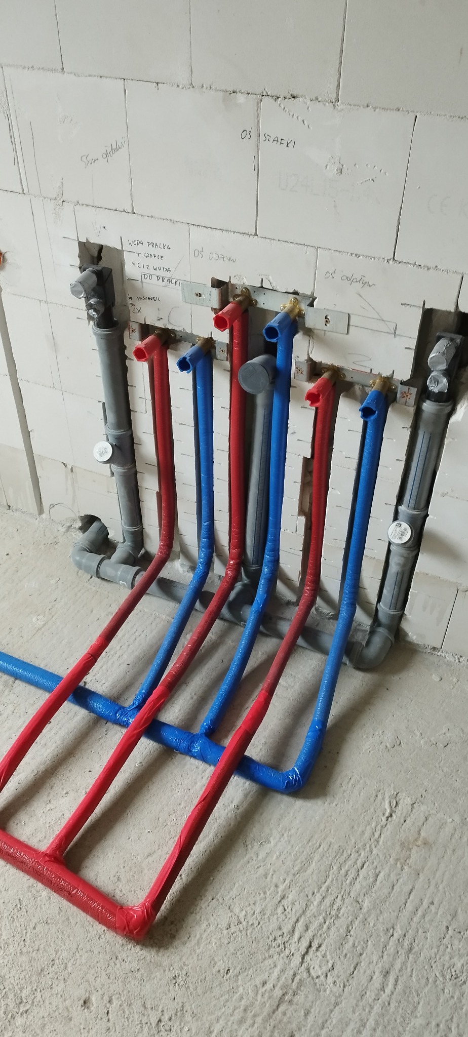 Instalacja wodna z rurami w kolorze czerwonym i niebieskim, zabezpieczonymi folią, zamontowana przy ścianie z bloczków. Widoczne napisy na ścianie.