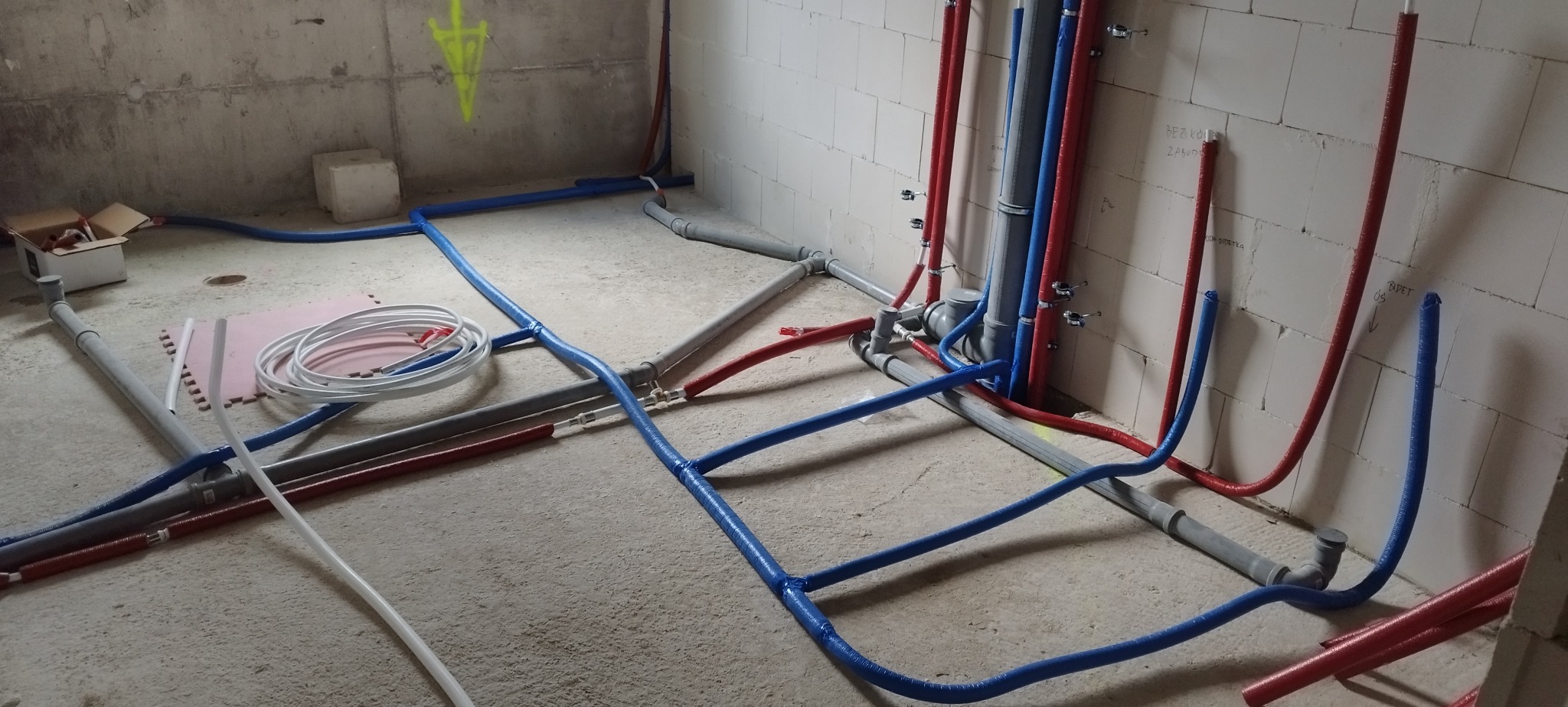 Instalacja wodna i kanalizacyjna w surowym wnętrzu. Widoczne rury w kolorach: niebieskim, czerwonym i szarym, prowadzone po podłodze i wzdłuż ścian z betonu komórkowego.