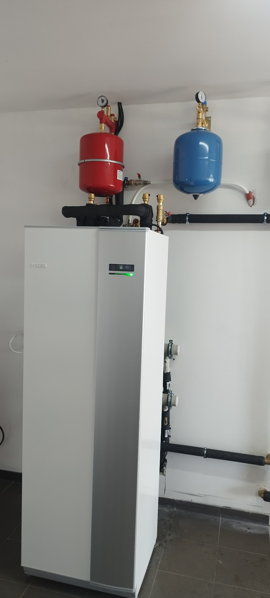 Biały piec CO marki Nibe z czerwonym i niebieskim zbiornikiem wyrównawczym, zamontowany w pomieszczeniu z szarą podłogą i białymi ścianami.