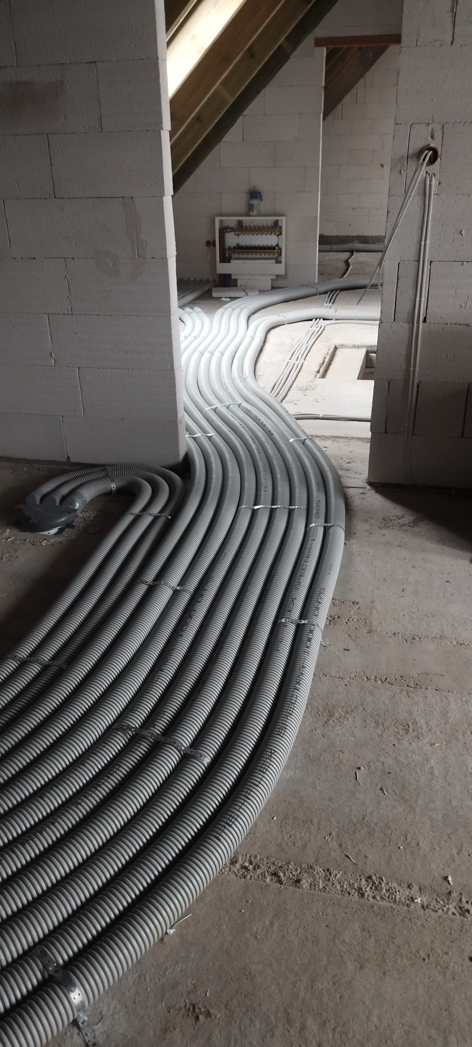 Szare rury karbowane ułożone równolegle na podłodze w surowym wnętrzu, prowadzące do rozdzielnicy na ścianie z białych bloczków. Widoczne mocowania rur.