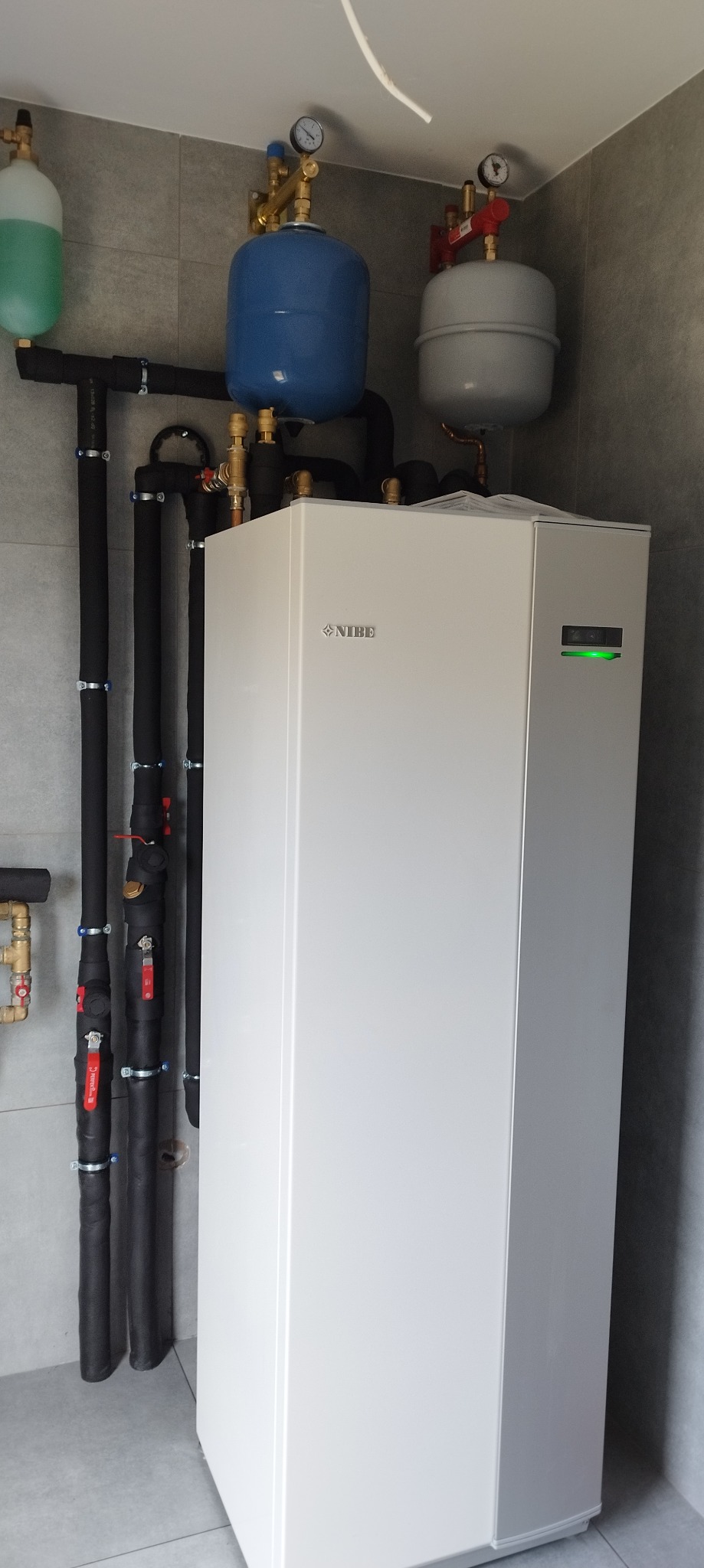 Nowoczesny, biały piec CO marki Nibe z zieloną diodą, zbiorniki wyrównawcze w kolorach niebieskim i szarym, czarne rury instalacji grzewczej na tle szarych płytek.