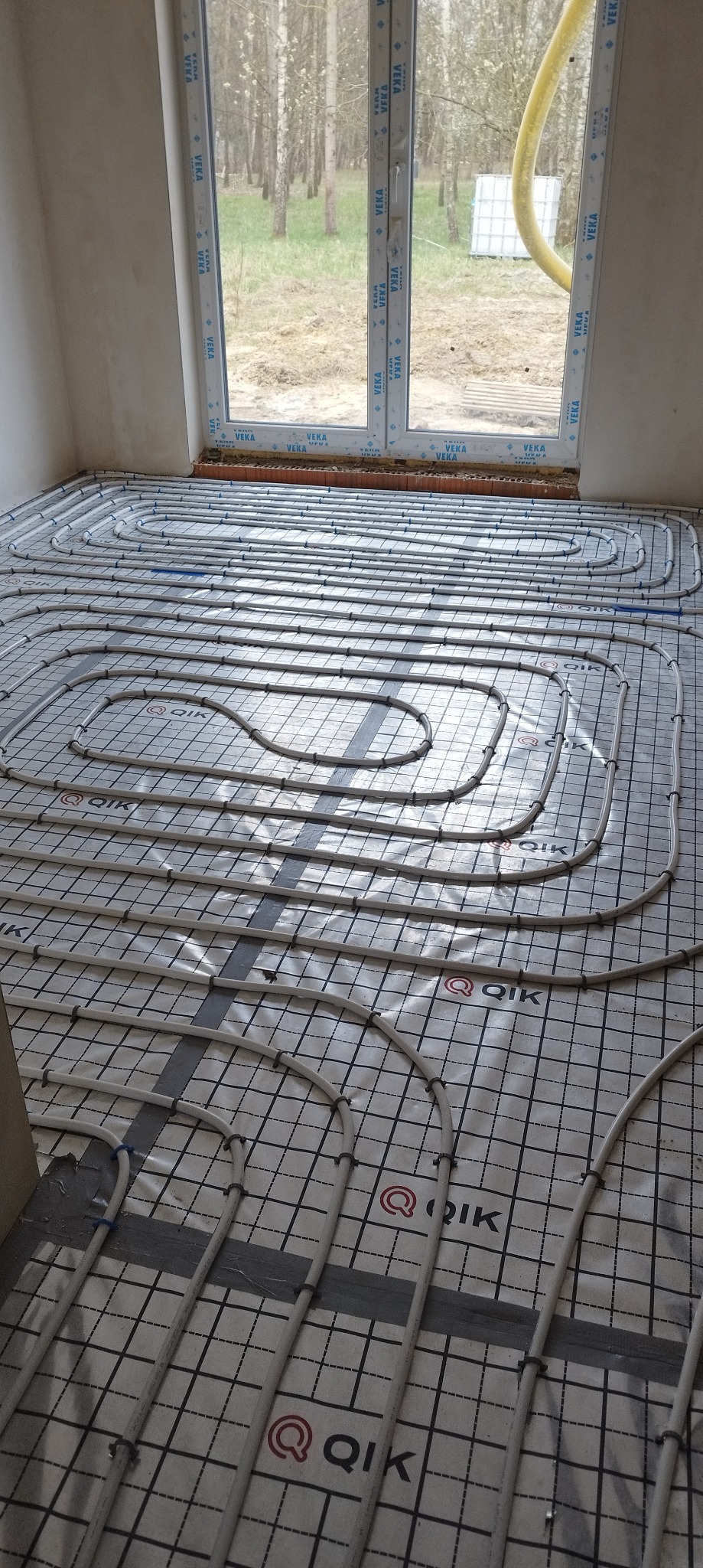 Białe rury ogrzewania podłogowego ułożone na folii z nadrukiem QIK, widoczne przez okno drzewa.