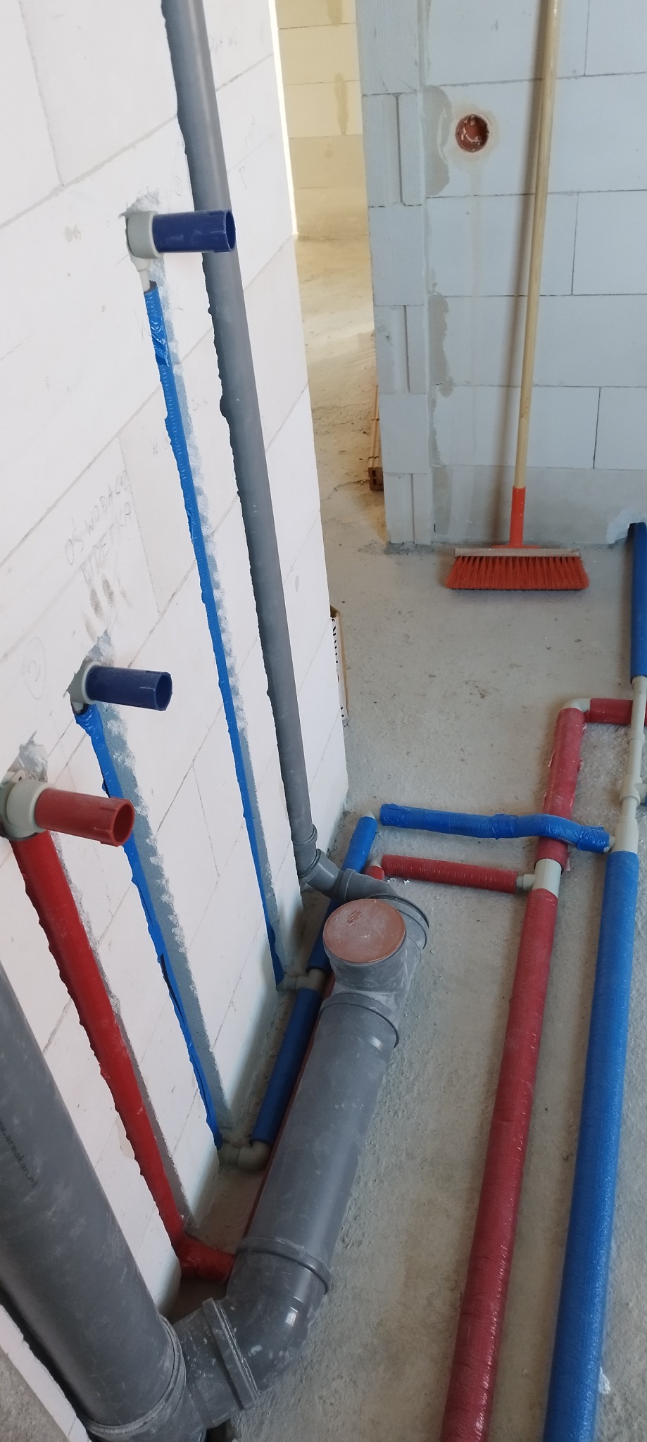 Instalacja wodna w surowym wnętrzu: rury z tworzywa sztucznego w kolorach czerwonym, niebieskim i szarym, częściowo ukryte w ścianie z betonu komórkowego.