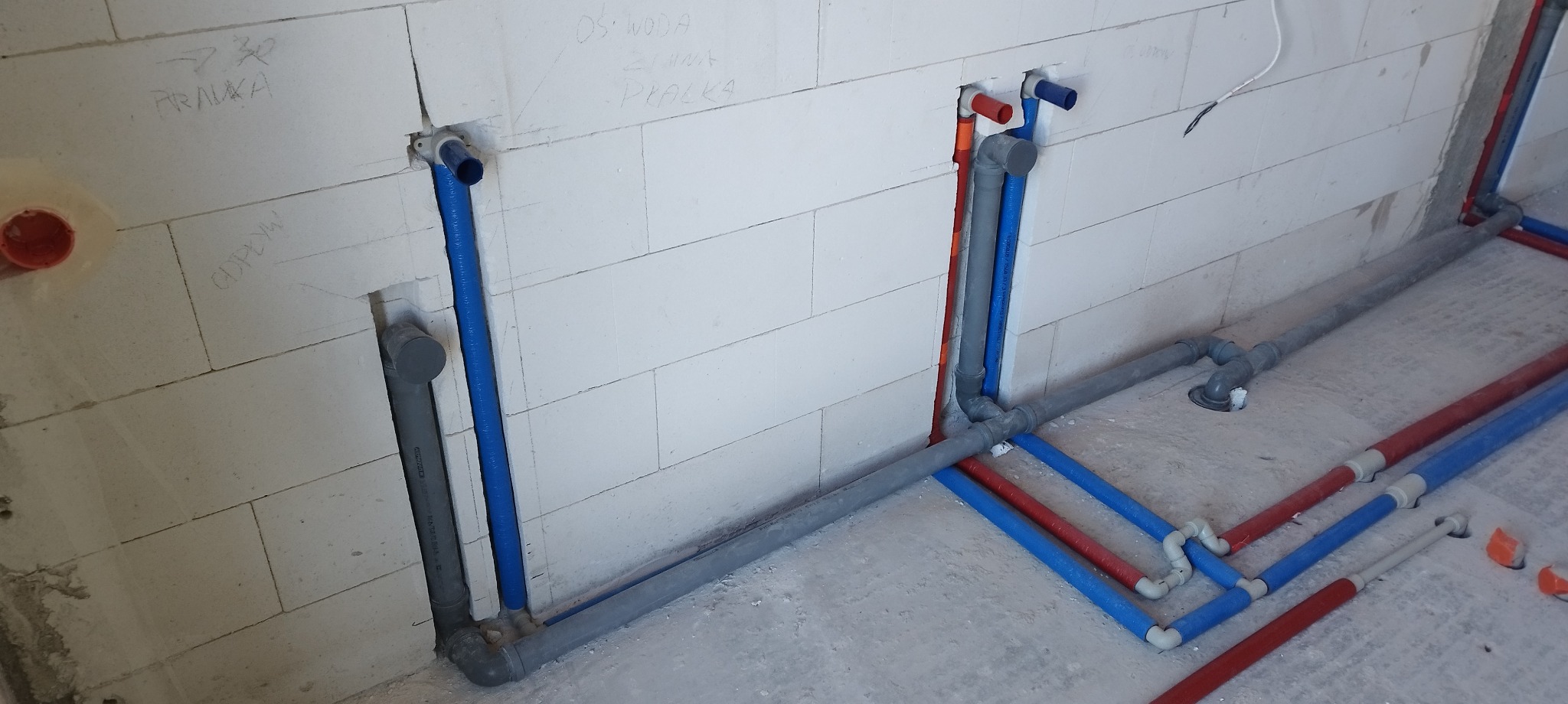 Instalacja wodno-kanalizacyjna w trakcie montażu, widoczne rury w kolorze niebieskim, czerwonym i szarym, prowadzone w ścianach z białego betonu komórkowego.