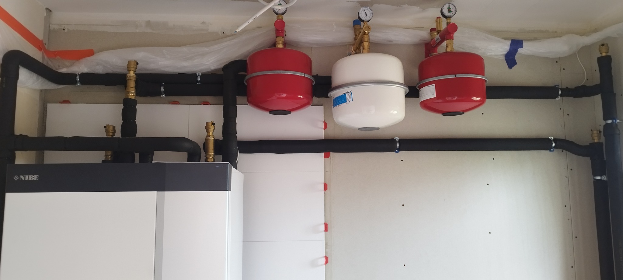 Instalacja CO z czarnymi rurami, czerwonymi i białym zbiornikiem wyrównawczym, manometrami, zamontowana na białej ścianie. Widoczny piec Nibe.