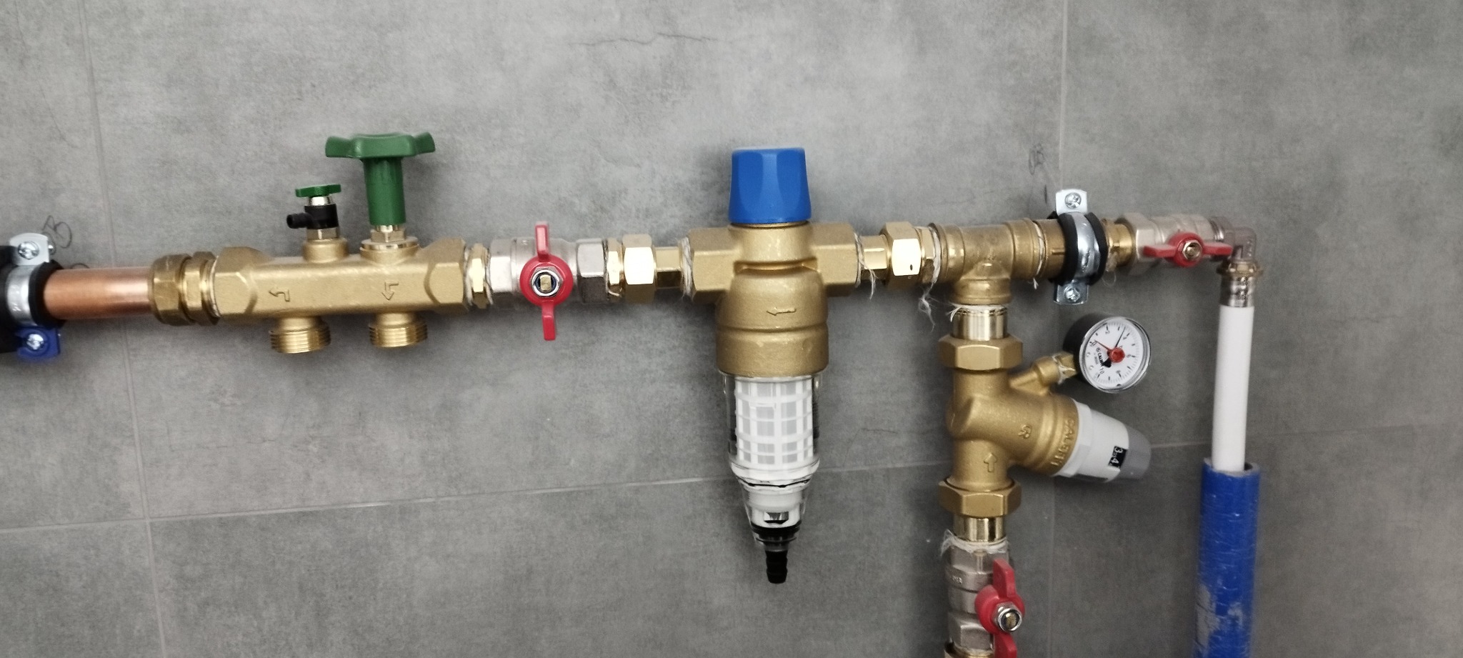 Kompletna instalacja wodna z filtrem, zaworami i manometrem, zamontowana na szarej, kafelkowanej ścianie. Widoczne miedziane i plastikowe rury oraz elementy z mosiądzu.