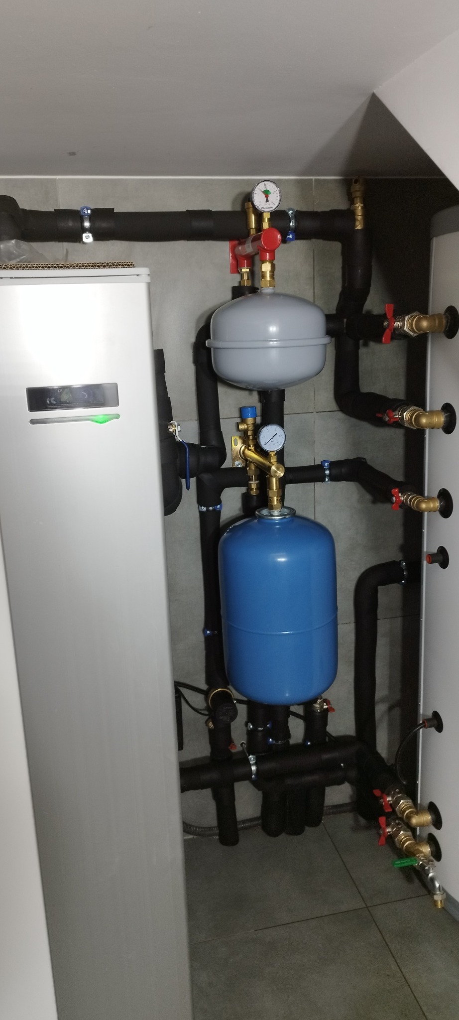 Nowoczesna kotłownia z szarym piecem i niebieskim zbiornikiem wyrównawczym, czarne rury z zaworami i manometrami na tle szarych płytek.