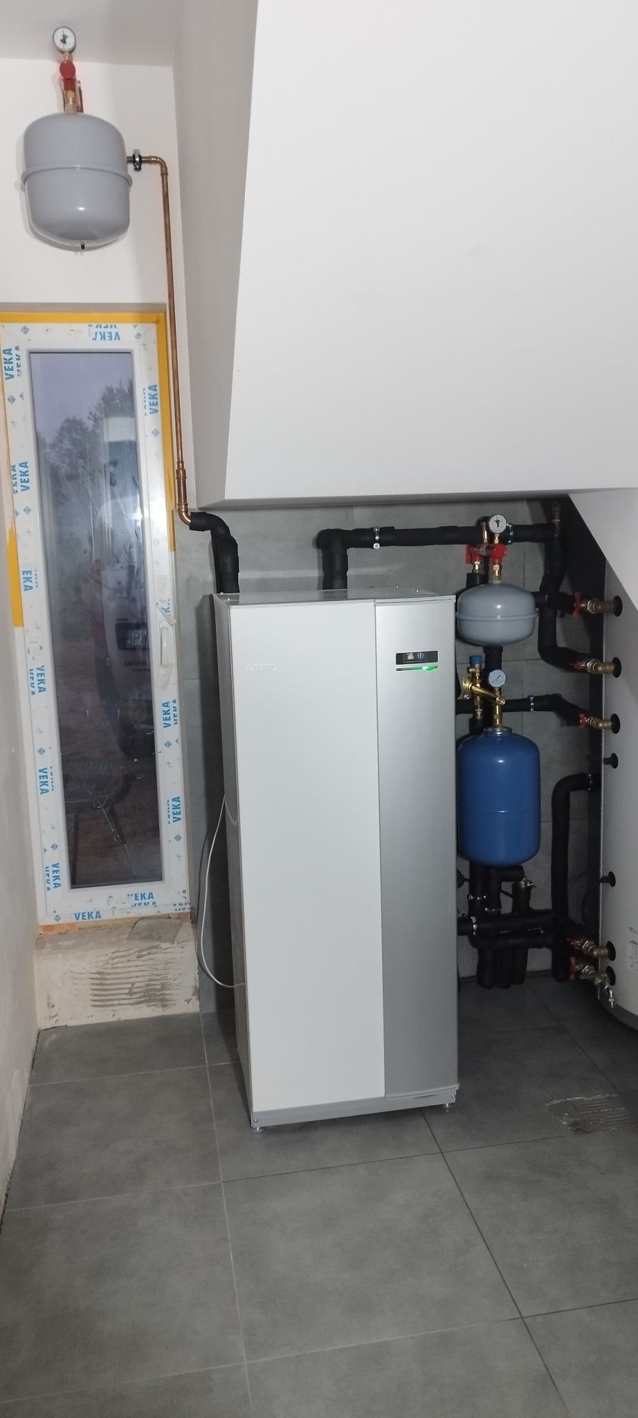 Nowoczesna instalacja grzewcza z szarym piecem i niebieskim zbiornikiem wyrównawczym, połączona rurami w pomieszczeniu z szarą podłogą i oknem.