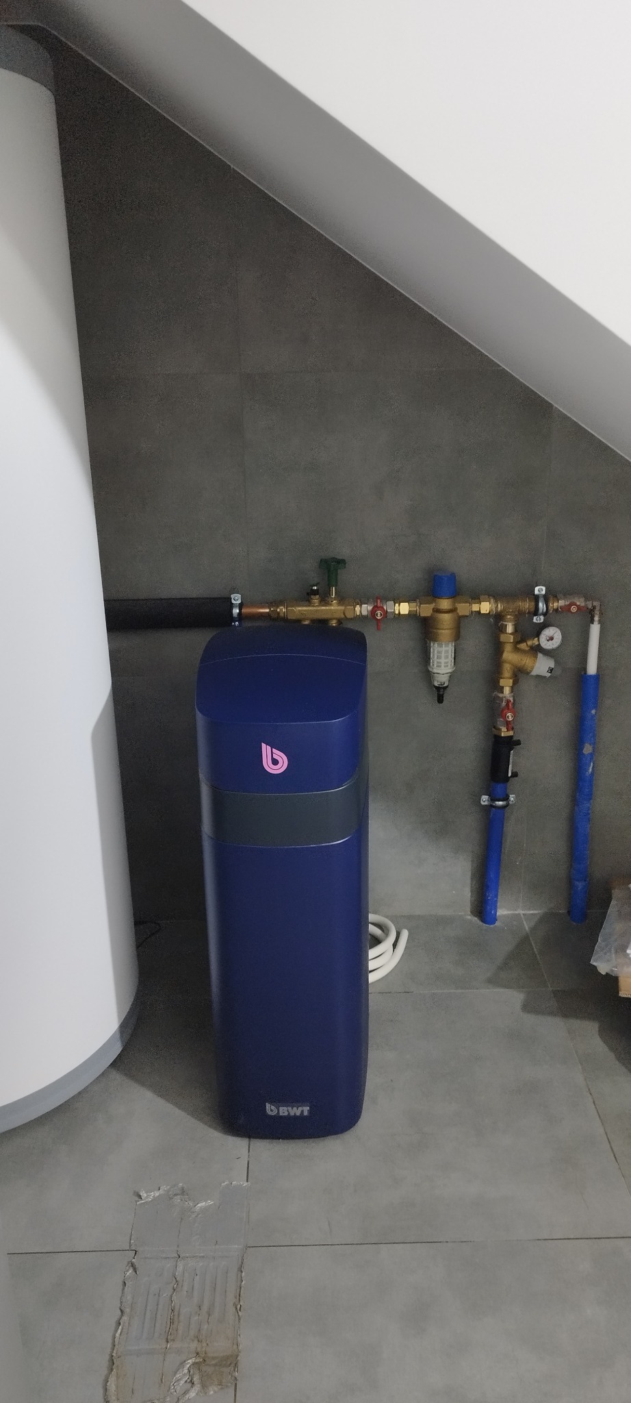 Niebieski zmiękczacz wody BWT, pionowo, obok białego bojlera w pomieszczeniu z szarą płytką. Widoczne rury, zawory i filtry instalacji wodnej. Ujęcie w skośnym pomieszczeniu.