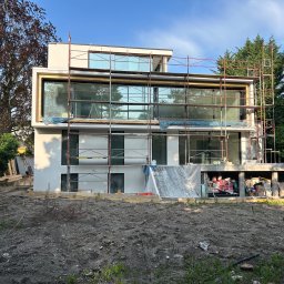 SOLICH TEAM SPÓŁKA Z OGRANICZONĄ ODPOWIEDZIALNOŚCIĄ - Nowoczesny dom w trakcie budowy, widoczne rusztowania i duże okna. Elementy wykończeniowe w jasnych barwach. Na pierwszym planie teren przygotowany pod ogród.