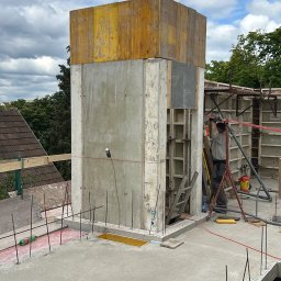 SOLICH TEAM SPÓŁKA Z OGRANICZONĄ ODPOWIEDZIALNOŚCIĄ - Wznoszenie betonowej konstrukcji z żółtym szalunkiem na budowie. Widoczny pracownik, zbrojenia i betonowa płyta. Ujęcie z góry, w tle zieleń i fragmenty zabudowy.