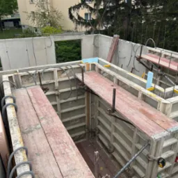 Szare szalunki fundamentów z deskami pomostowymi, widok z góry na budowę, z zielenią w tle i elementami instalacji. W tle budynek mieszkalny.