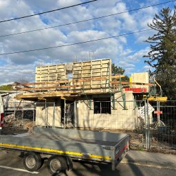 SOLICH TEAM SPÓŁKA Z OGRANICZONĄ ODPOWIEDZIALNOŚCIĄ - Wznoszona ściana domu z betonu komórkowego, widoczne szalunki i zbrojenia. Na pierwszym planie przyczepka i częściowo widoczny samochód dostawczy.