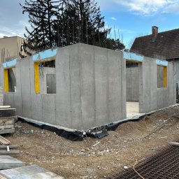 SOLICH TEAM SPÓŁKA Z OGRANICZONĄ ODPOWIEDZIALNOŚCIĄ - Surowy stan ścian parteru domu z betonu komórkowego, widoczne żółte ramy okienne oraz wystające pręty zbrojeniowe, teren budowy z elementami konstrukcyjnymi.