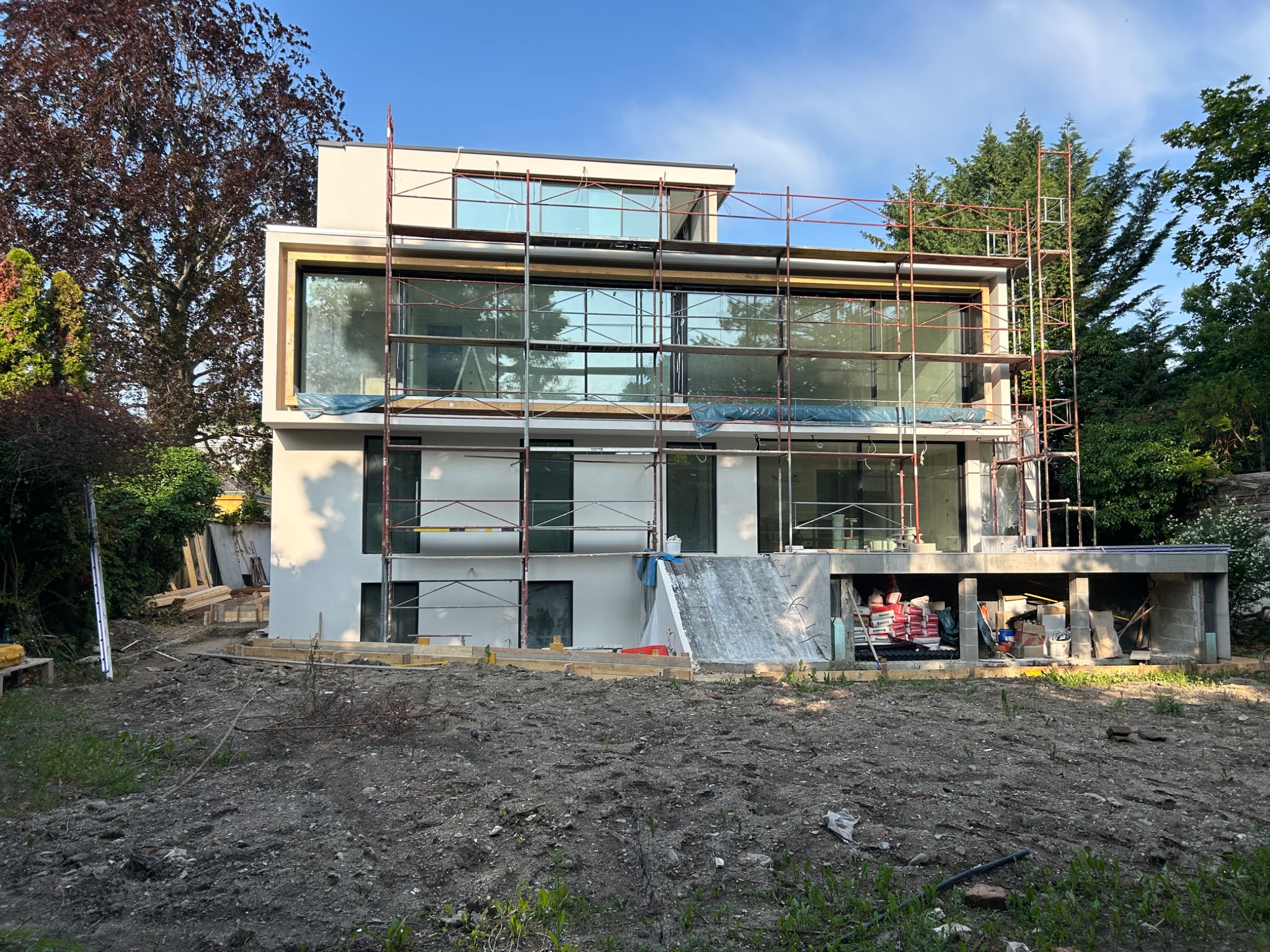 Nowoczesny dom w trakcie budowy, widoczne rusztowania i duże okna. Elementy wykończeniowe w jasnych barwach. Na pierwszym planie teren przygotowany pod ogród.