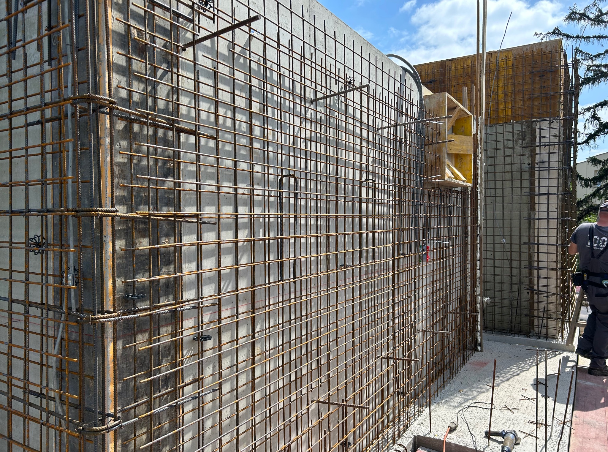 Zbrojenie pionowych ścian fundamentowych z betonu, widok z boku, zbrojenie w formie siatki z prętów stalowych, szalunki, fragment sylwetki pracownika w tle.