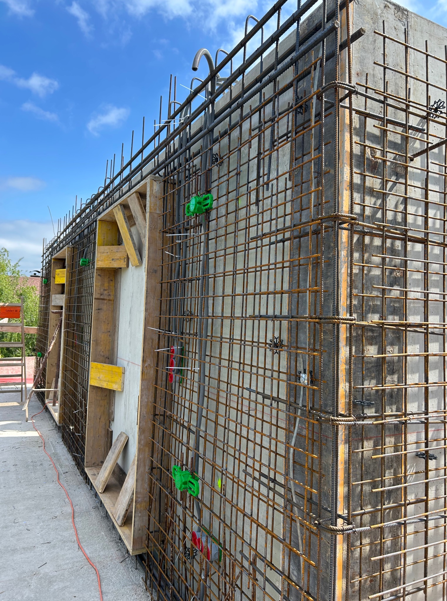 Zbrojenie pionowej ściany szalunkowej z widocznymi elementami drewnianymi i plastikowymi łącznikami, na tle błękitnego nieba z chmurami.