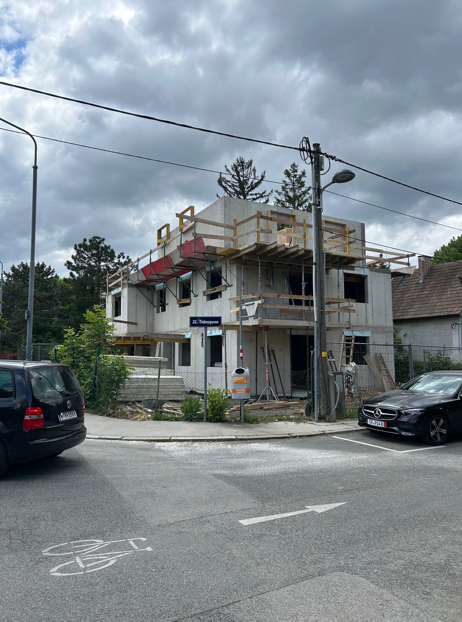 Surowy stan domu murowanego w trakcie budowy, widok z ulicy Thönygasse z samochodami w kadrze i pochmurnym niebem w tle. Widoczne rusztowania i materiały budowlane.