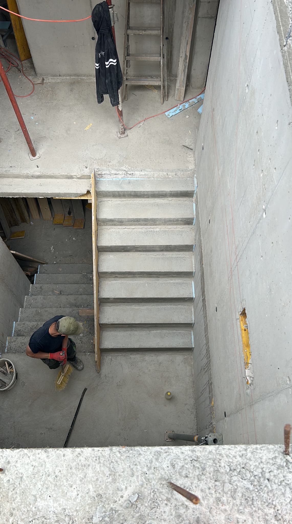 Betonowe schody w surowym stanie, widok z góry na pracownika z miotłą, drabina i odzież robocza w tle, surowy beton, oświetlenie z góry.