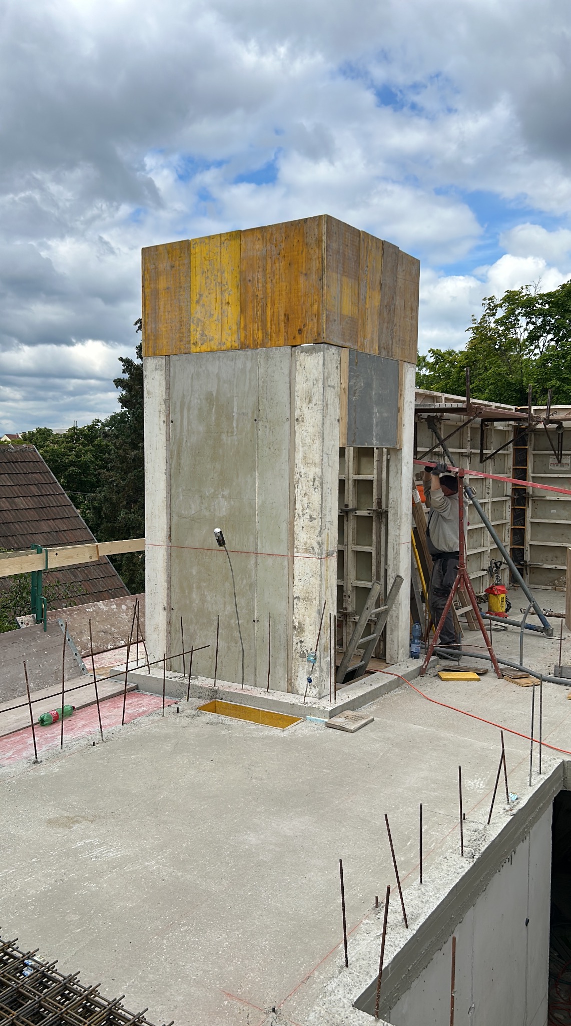 Wznoszenie betonowej konstrukcji z żółtym szalunkiem na budowie. Widoczny pracownik, zbrojenia i betonowa płyta. Ujęcie z góry, w tle zieleń i fragmenty zabudowy.