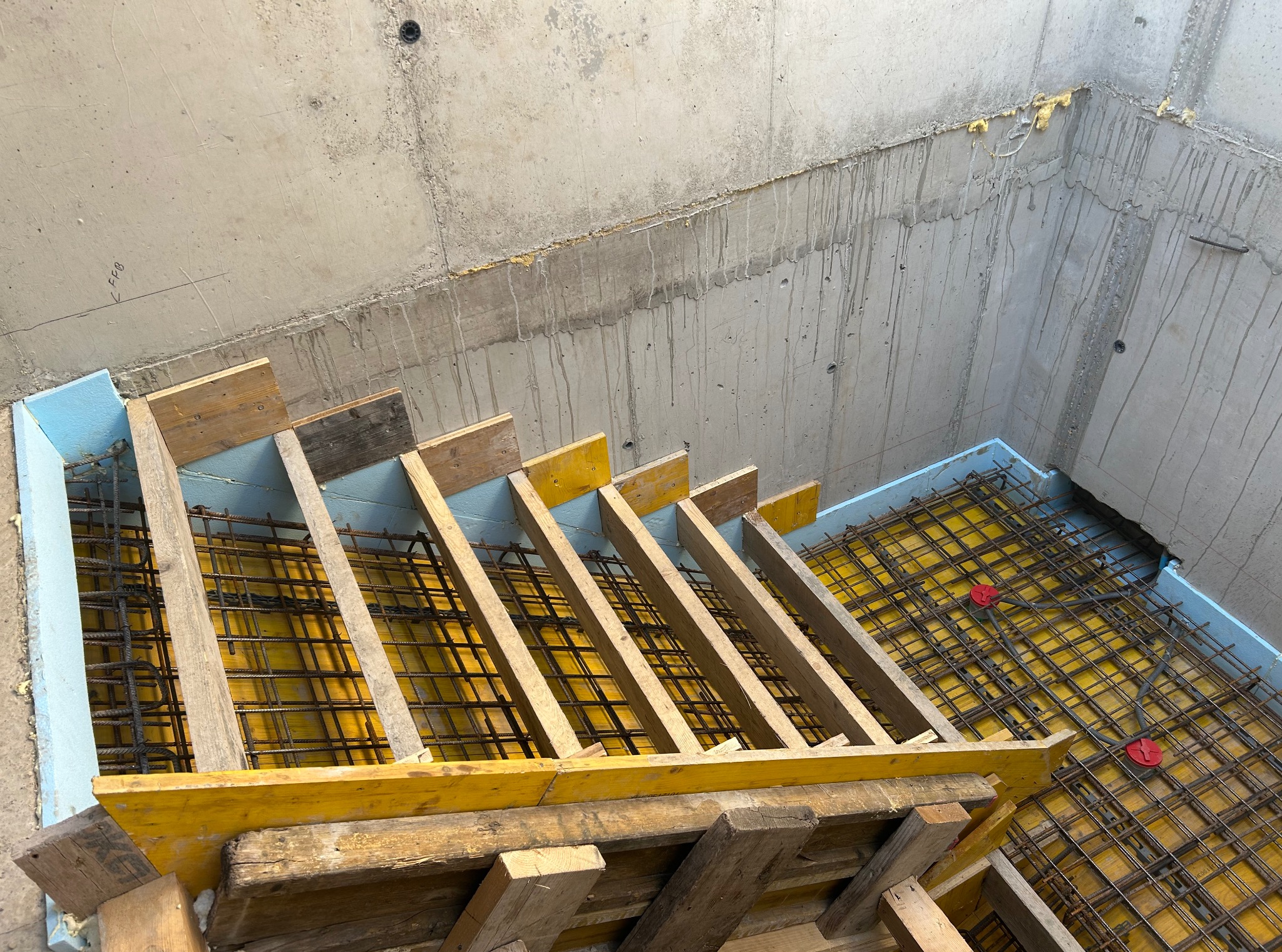 Zbrojenie fundamentów z szalunkiem i izolacją termiczną, widok z góry. Żółta mata izolacyjna i stalowe pręty tworzą siatkę. Betonowe ściany w tle.