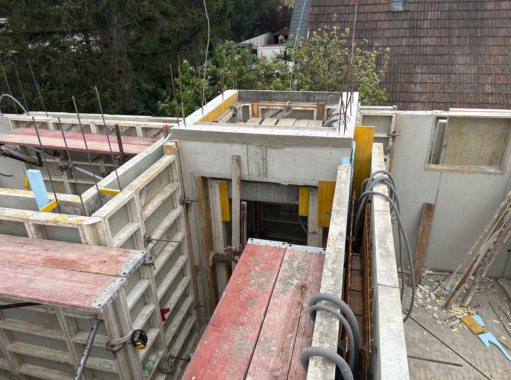 Szare szalunki fundamentów z drewnianymi belkami i elementami zbrojenia, widok z góry na plac budowy z zielenią w tle.